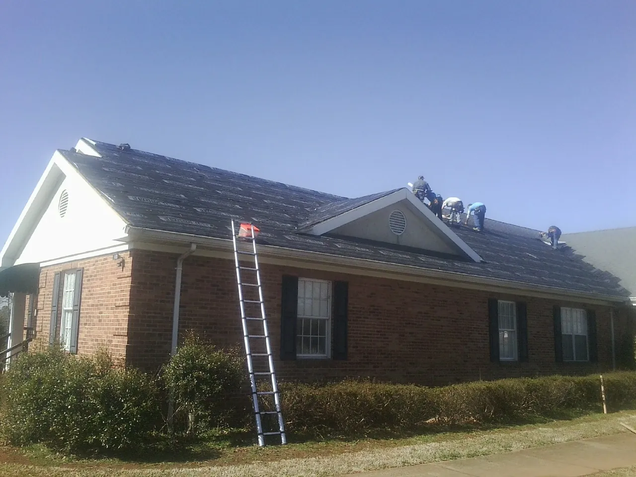 Crew tearing shingles off a brick ranch roof with a ladder set — Emergency Roof Repair work by our crew in Holland Patent