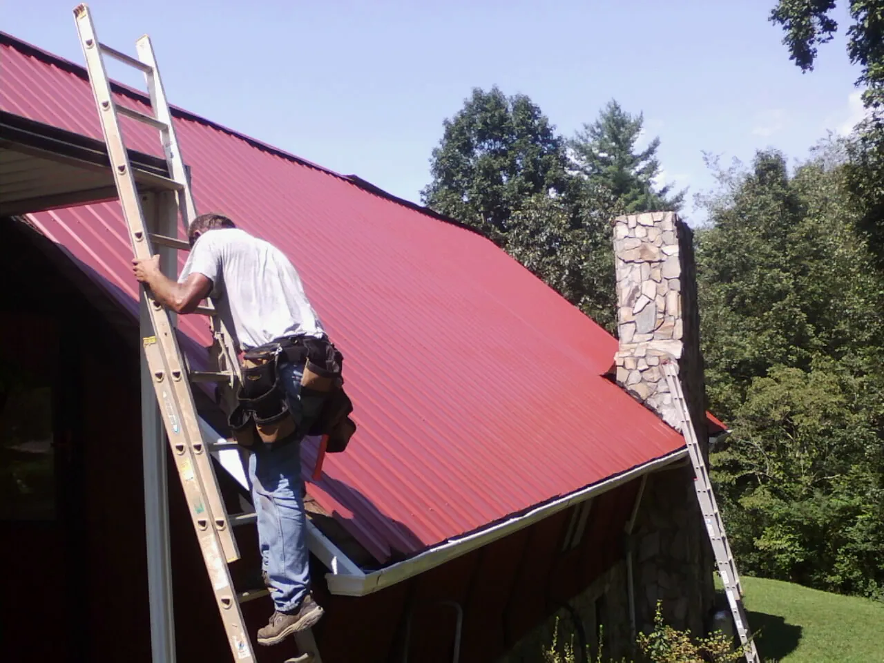 Roofer on a ladder beside a red metal roof and stone chimney — Soffit & Fascia Repair by Roofing Brothers in Holland Patent