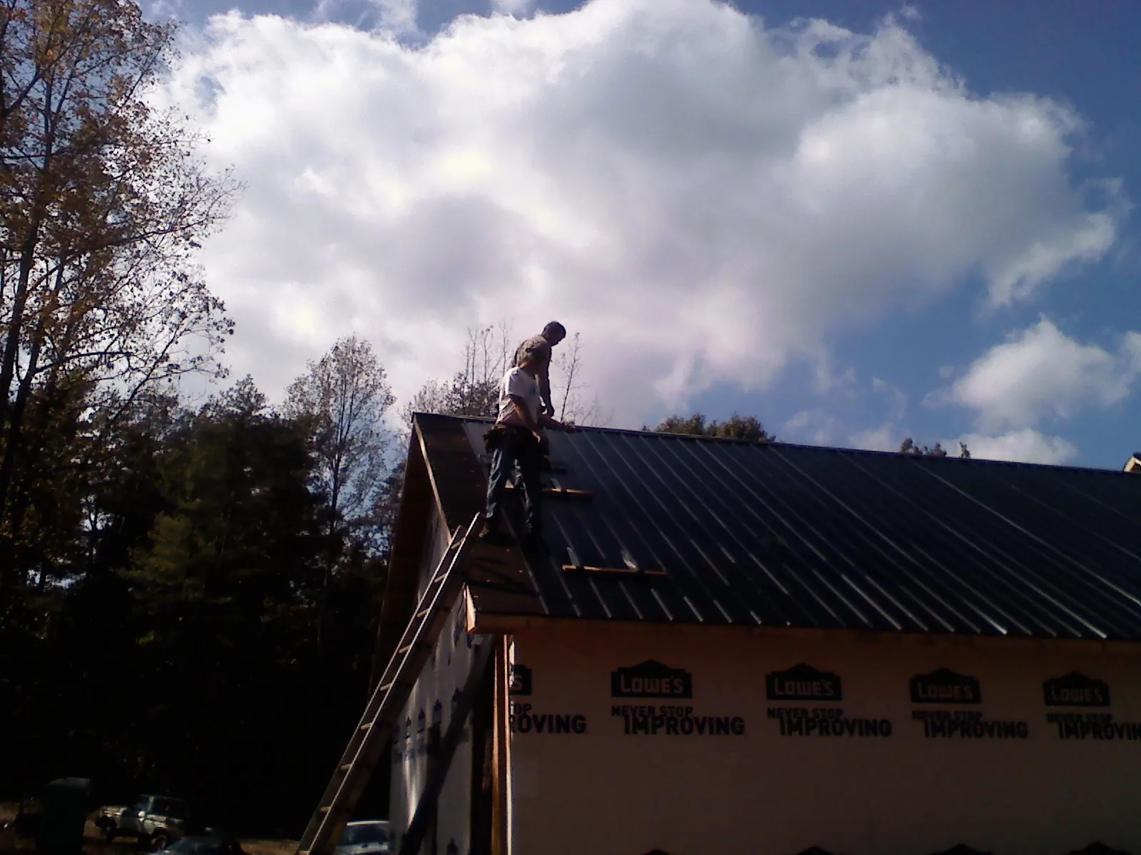 Roofer fastening dark metal panels high against a cloudy sky, from a Roof Inspection job in Holland Patent